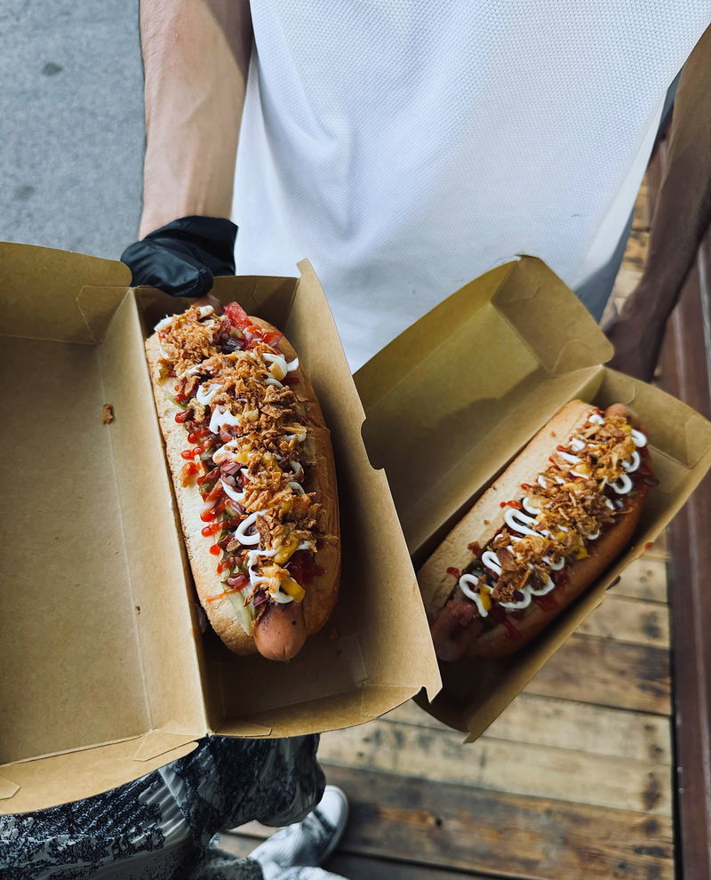 Two hot dogs in kraft boxes held by a member of staff