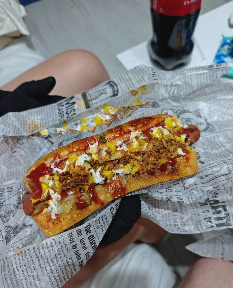 A hot dog in hand with a bottle of cola on the table