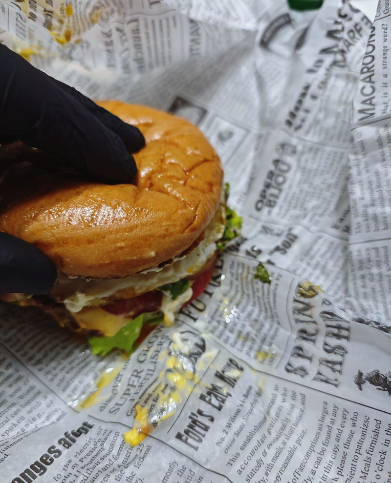 A gloved hand holding a burger on newspaper-print wrap — gloves come with every order