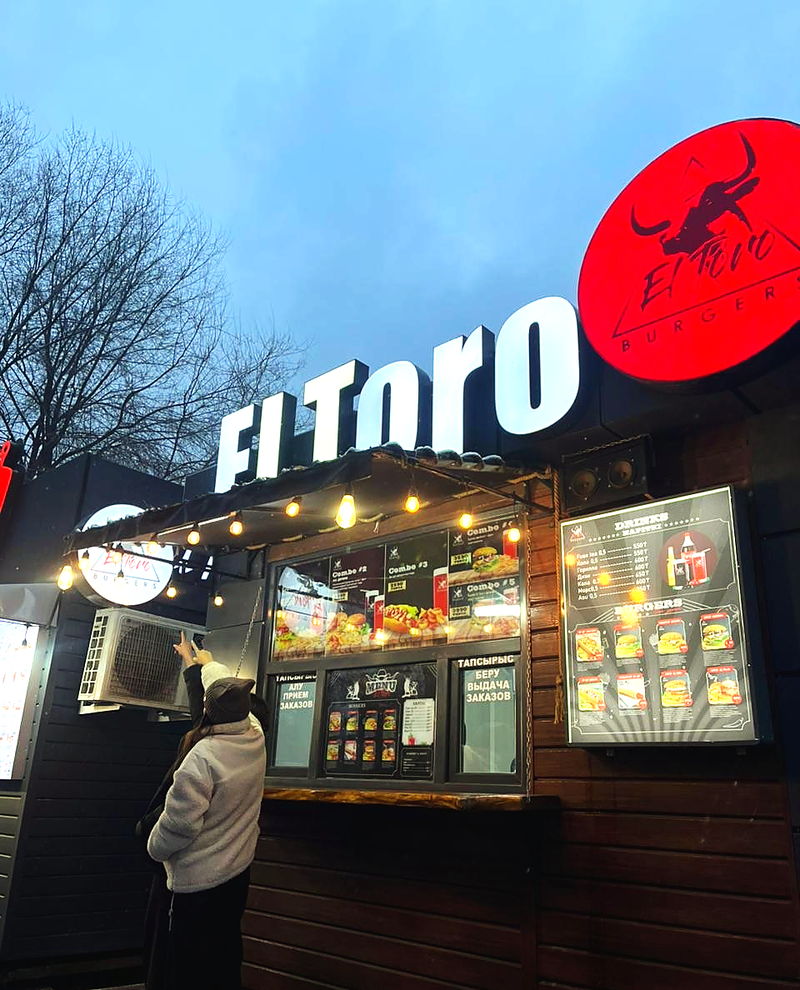 Павильон El Toro Burgers в сумерках: белые объёмные буквы «El Toro», красный круглый лайтбокс с быком и гирлянда ламп над окном выдачи