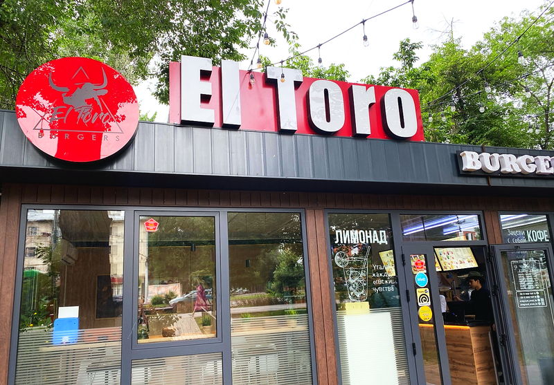 The El Toro Burgers frontage on Abay Avenue by day: a red fascia with white letters, the round bull logo and a string of bulbs