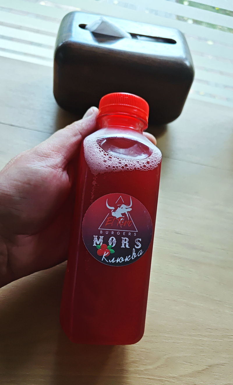 A bottle of El Toro’s house cranberry mors, its round “MORS Клюква” label held up by a guest